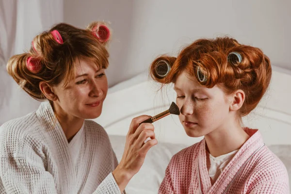 Família feliz. Jovem mãe bonita faz maquiagem de sua linda filha na cama no quarto. A filha ruiva com caracóis na cabeça está a rir. . — Fotografia de Stock