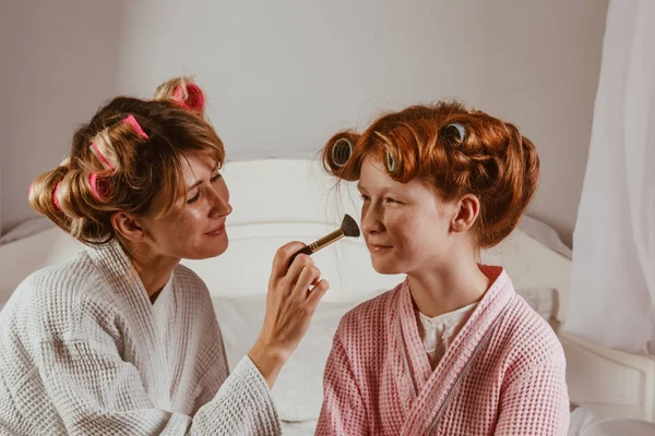 Счастливая семья. Молодая красивая мать делает макияж своей прекрасной дочери на кровати в спальне. Рыжая дочь с бигудями на голове смеется . — стоковое фото