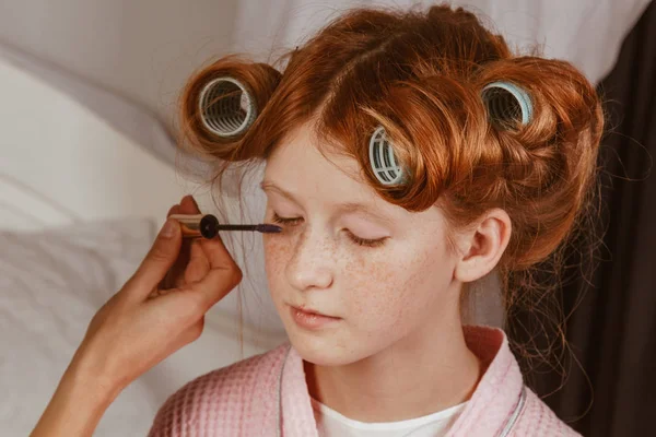 Família feliz. Jovem mãe bonita faz maquiagem de sua linda filha na cama no quarto. A filha ruiva com caracóis na cabeça está a rir. . — Fotografia de Stock