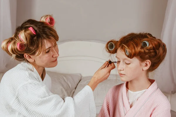 Família feliz. Jovem mãe bonita faz maquiagem de sua linda filha na cama no quarto. A filha ruiva com caracóis na cabeça está a rir. . — Fotografia de Stock