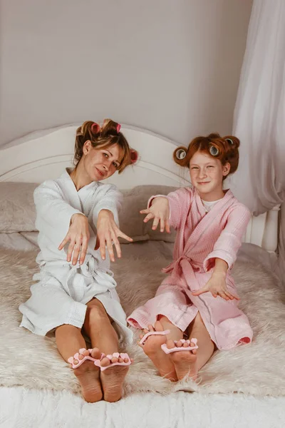 Boldog szerető családot. Anya és lánya manikűröznek, pedikűröznek, sminkelnek és szórakoznak. Anya és a kislány fürdőköpenyben és hajcsavaróval a fejükön.. — Stock Fotó