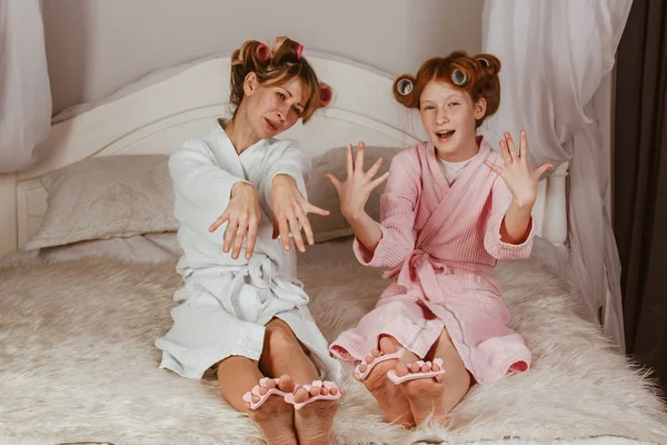 Happy loving family. Mom and daughter do manicures, pedicures, do makeup and have fun. Mom and little girl in bathrobes and with curlers on their heads.