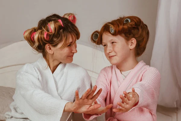 Feliz familia amorosa. Mamá e hija hacen manicuras, pedicuras, maquillan y se divierten. Mamá y niña con albornoces y rizadores en la cabeza . — Foto de Stock