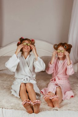 Tatil günü evde. Anne ve kızı evde gözlerinde salatalık maskesi yapıyor. Güzellik salonu konsepti. Güzel bir yüz. Kadın ve mutlu bebek. Bigudili kızlar.