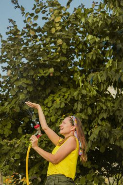 Genç ve güzel bir kız bahçede, çiçekleri hortumla sularken eğleniyor. En sevdiğim hobimi yaparken gülümsüyorum..