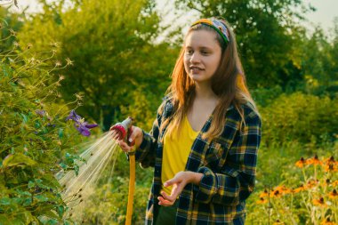 Genç ve güzel bir kız bahçede, çiçekleri hortumla sularken eğleniyor. En sevdiğim hobimi yaparken gülümsüyorum..