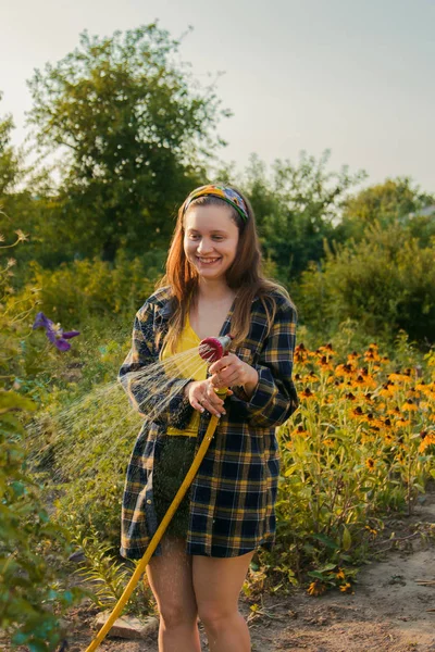 Jovem menina bonita se divertindo no jardim regando plantas com uma mangueira. Sorrindo enquanto toma um hobby favorito . — Fotografia de Stock
