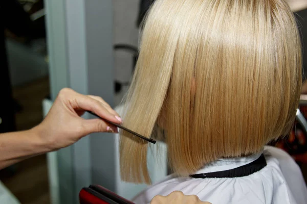 Účes. Atraktivní blondýna s krátkými vlasy dělá účes s elektrickým žehličkou. Narovnávání vlasů šedé pozadí. detailní záběr. Fotografie makra. — Stock fotografie