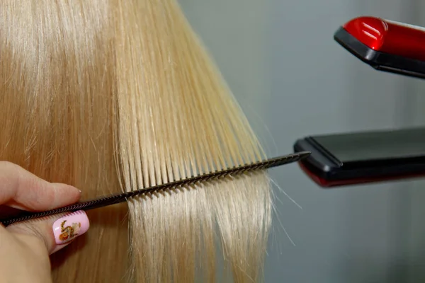 Účes. Atraktivní blondýna s krátkými vlasy dělá účes s elektrickým žehličkou. Narovnávání vlasů šedé pozadí. detailní záběr. Fotografie makra. — Stock fotografie
