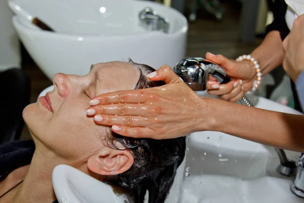 Nádherná veselá starší žena si užívá masáž hlavy a zároveň si myje vlasy profesionálním kadeřníkem. Krása péče, účes, móda, životní styl okouzlující koncept. Kapky vody a šampon pěny. — Stock fotografie