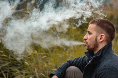 Sakallı bir adam gölde oturur ve elektronik sigara içer. Duman üflüyor. Doğayla bütünlük kavramı. Suyun yanında dinlen. Vape, Erkek Vaping. dudak ve duman, bir sürü duman..