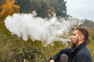Sakallı bir adam gölde oturur ve elektronik sigara içer. Duman üflüyor. Doğayla bütünlük kavramı. Suyun yanında dinlen. Vape, Erkek Vaping. dudak ve duman, bir sürü duman..