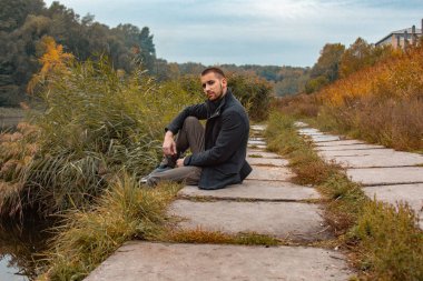 Sakallı bir adam gölde oturur ve elektronik sigara içer. Duman üflüyor. Doğayla bütünlük kavramı. Suyun yanında dinlen. Vape, Erkek Vaping. dudak ve duman, bir sürü duman..