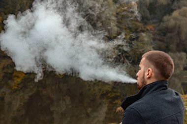 Sakallı bir adam gölde oturur ve elektronik sigara içer. Duman üflüyor. Doğayla bütünlük kavramı. Suyun yanında dinlen. Vape, Erkek Vaping. dudak ve duman, bir sürü duman..