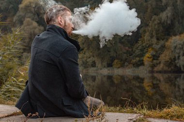 Sakallı bir adam gölde oturur ve elektronik sigara içer. Duman üflüyor. Doğayla bütünlük kavramı. Suyun yanında dinlen. Vape, Erkek Vaping. dudak ve duman, bir sürü duman..