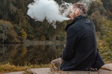Sakallı bir adam gölde oturur ve elektronik sigara içer. Duman üflüyor. Doğayla bütünlük kavramı. Suyun yanında dinlen. Vape, Erkek Vaping. dudak ve duman, bir sürü duman..