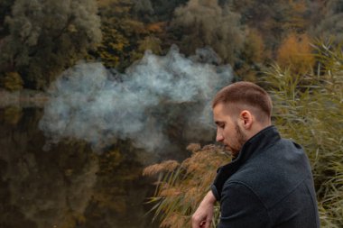 Sakallı bir adam gölde oturur ve elektronik sigara içer. Duman üflüyor. Doğayla bütünlük kavramı. Suyun yanında dinlen. Vape, Erkek Vaping. dudak ve duman, bir sürü duman..