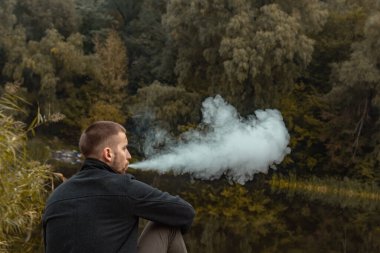 Sakallı bir adam gölde oturur ve elektronik sigara içer. Duman üflüyor. Doğayla bütünlük kavramı. Suyun yanında dinlen. Vape, Erkek Vaping. dudak ve duman, bir sürü duman..