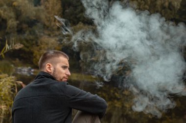 Sakallı bir adam gölde oturur ve elektronik sigara içer. Duman üflüyor. Doğayla bütünlük kavramı. Suyun yanında dinlen. Vape, Erkek Vaping. dudak ve duman, bir sürü duman..