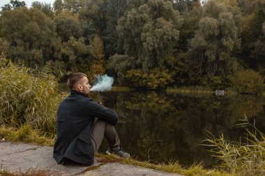 Sakallı bir adam gölde oturur ve elektronik sigara içer. Duman üflüyor. Doğayla bütünlük kavramı. Suyun yanında dinlen. Vape, Erkek Vaping. dudak ve duman, bir sürü duman..