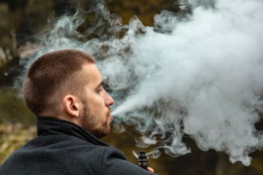 Sakallı bir adam gölde oturur ve elektronik sigara içer. Duman üflüyor. Doğayla bütünlük kavramı. Suyun yanında dinlen. Vape, Erkek Vaping. dudak ve duman, bir sürü duman..