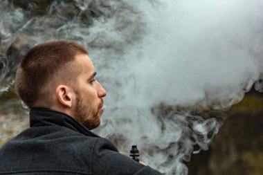 Sakallı bir adam gölde oturur ve elektronik sigara içer. Duman üflüyor. Doğayla bütünlük kavramı. Suyun yanında dinlen. Vape, Erkek Vaping. dudak ve duman, bir sürü duman..