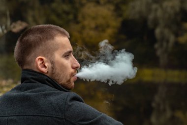 Sakallı bir adam gölde oturur ve elektronik sigara içer. Duman üflüyor. Doğayla bütünlük kavramı. Suyun yanında dinlen. Vape, Erkek Vaping. dudak ve duman, bir sürü duman..