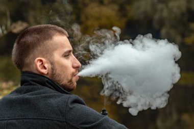 Sakallı bir adam gölde oturur ve elektronik sigara içer. Duman üflüyor. Doğayla bütünlük kavramı. Suyun yanında dinlen. Vape, Erkek Vaping. dudak ve duman, bir sürü duman..