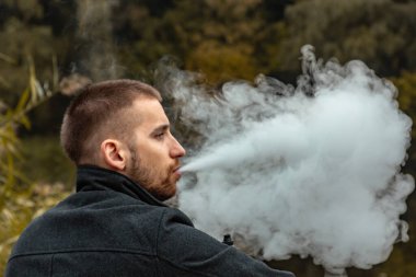Sakallı bir adam gölde oturur ve elektronik sigara içer. Duman üflüyor. Doğayla bütünlük kavramı. Suyun yanında dinlen. Vape, Erkek Vaping. dudak ve duman, bir sürü duman..