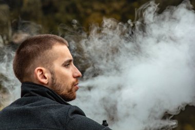 Sakallı bir adam gölde oturur ve elektronik sigara içer. Duman üflüyor. Doğayla bütünlük kavramı. Suyun yanında dinlen. Vape, Erkek Vaping. dudak ve duman, bir sürü duman..