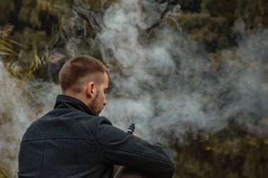 Sakallı bir adam gölde oturur ve elektronik sigara içer. Duman üflüyor. Doğayla bütünlük kavramı. Suyun yanında dinlen. Vape, Erkek Vaping. dudak ve duman, bir sürü duman..