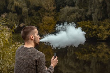 Sakallı bir adam gölde oturur ve elektronik sigara içer. Duman üflüyor. Doğayla bütünlük kavramı. Suyun yanında dinlen. Vape, Erkek Vaping. dudak ve duman, bir sürü duman..
