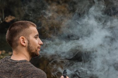 Sakallı bir adam gölde oturur ve elektronik sigara içer. Duman üflüyor. Doğayla bütünlük kavramı. Suyun yanında dinlen. Vape, Erkek Vaping. dudak ve duman, bir sürü duman..