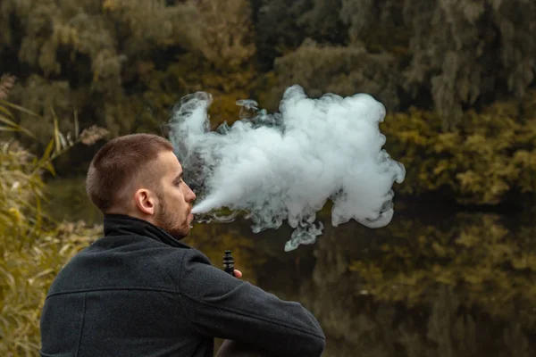 Sakallı bir adam gölde oturur ve elektronik sigara içer. Duman üflüyor. Doğayla bütünlük kavramı. Suyun yanında dinlen. Vape, Erkek Vaping. dudak ve duman, bir sürü duman..