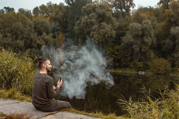 Un tipo con barba se sienta en el lago y fuma un cigarrillo electrónico. Sopla bocanadas de humo. El concepto de unidad con la naturaleza. Descansa cerca del agua. Vape, Man Vaping. labios y humo, mucho humo . —  Fotos de Stock