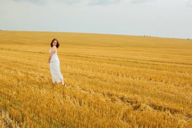Güzel bir açık hava eğlencesi. Buğday tarlasının ortasında güzel, kızıl saçlı bir kadın eğleniyor. Yaz manzarası, güzel hava. Güneşli ve bulutlu rüzgarlı bir gün. Beyaz pamuklu elbise, eko tarzı.