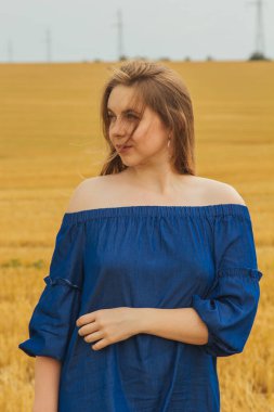 Buğday tarlasının ortasında güzel sarışın bir kız. Kameraya bakarak poz veriyorsun. Yaz manzarası, güzel hava. Mavi pamuklu elbise, eko tarzı. Güzel çizgili şapka