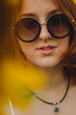 Beyaz elbiseli, gözlüklü, güzel, seksi, kızıl saçlı bir kızın portresi. Ayçiçeklerinde yürür. Zevk duygusu, özgürlük konsepti, yaşam tarzı. Güneş gözlüğü reklamı