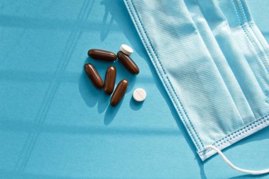 Vitaminler ve doktor maskesi, mavi arka planda taç virüsü koruma konsepti. Güneşten gelen gölgeler. Solunum maskesi ve haplar. Coronavirus, hastane veya çevre kirliliği yüzleri korur