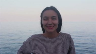 Mavi denizin arka planında güzel bir kızın portresi. Deniz ve plajların arka planında çekici bir kızın yakın plan portresi. Kameraya bakıyor, gülümsüyor ve elini selamlıyor..