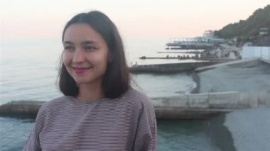 Mavi denizin arka planında güzel bir kızın portresi. Deniz ve plajların arka planında çekici bir kızın yakın plan portresi. Kameraya bakıyor, gülümsüyor ve elini selamlıyor..