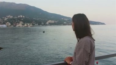 Mavi deniz ve dağların arka planında güzel bir kızın portresi. Deniz ve plajların arka planında çekici bir kızın yakın plan portresi. Kameraya bakıyor, gülümsüyor ve elini sallıyor..