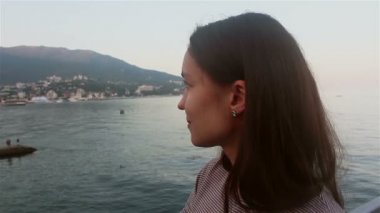 Mavi deniz ve dağların arka planında güzel bir kızın portresi. Deniz ve plajların arka planında çekici bir kızın yakın plan portresi. Kameraya bakıyor, gülümsüyor ve elini sallıyor..