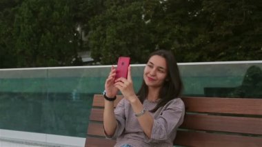 Genç ve güzel bir kız parktaki bankta otururken akıllı telefonuyla selfie çekiyor. Parktaki bir bankta rahatlayan ve akıllı telefonuyla selfie çeken çekici bir kızın yakın plan portresi..