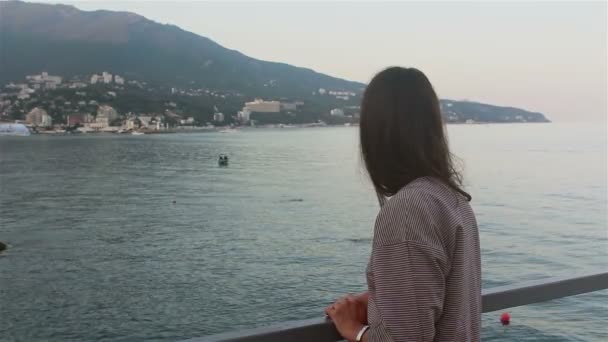 在蓝色的大海和群山的背景上描绘一个年轻漂亮的女孩 在大海和海滩的背景上 一位漂亮姑娘的近照 她看着相机 微笑着挥挥手致意 — 图库视频影像