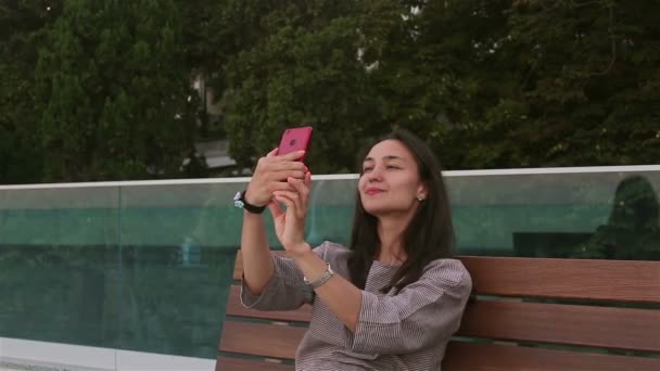年轻美丽的姑娘坐在公园的长椅上 一边用智能手机自私自利 一位年轻貌美的姑娘的特写 她正在公园的长椅上休息 还在智能手机上自拍 — 图库视频影像