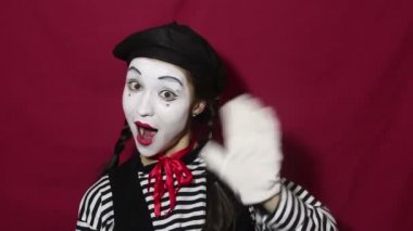 Genç bir pandomimci kızın yakın plan portresi. Güzel pandomimci kız gülümseyip elini sallıyor. Pandomimci kız müşterileri ağırlarken elini sallıyor. Kırmızı arkaplanda bir portre.