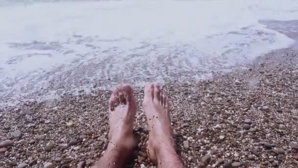 Vista de las piernas de un hombre acostado en la orilla del mar. Piernas masculinas sobre un fondo de mar azul.Un hombre disfruta de unas vacaciones en una playa de guijarros . — Vídeos de Stock