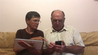 Çekici yetişkin kadın ve yetişkin adam tablet bilgisayar ve akıllı telefon üzerinde çalışıyorlar. Yaşlı bir çift evdeki kanepede oturup tablet bilgisayarda ve akıllı telefondan fotoğraf izleyip gülümsüyor ve aktif bir şekilde konuşuyor..