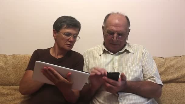 迷人的成年女性和成年男性在平板电脑和智能手机上工作 一对年长的夫妇坐在家中的沙发上 看着平板电脑和智能手机上的照片 微笑着 积极地交谈着 — 图库视频影像